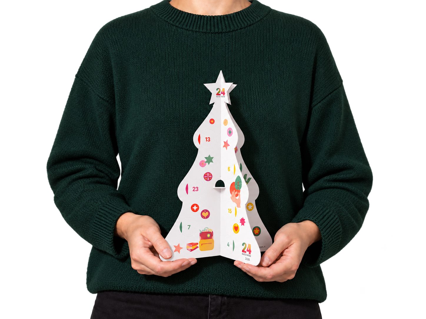 Der 24 GUTE TATEN Adventskalender 2026 als aufklappbarer Papier-Weihnachtsbaum, gehalten von einer Person im grünen Pullover