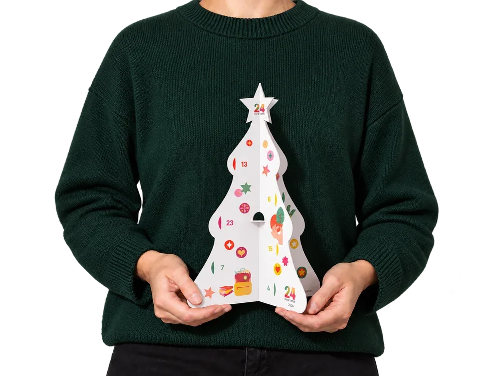 Der 24 GUTE TATEN Adventskalender 2026 als aufklappbarer Papier-Weihnachtsbaum, gehalten von einer Person im grünen Pullover
