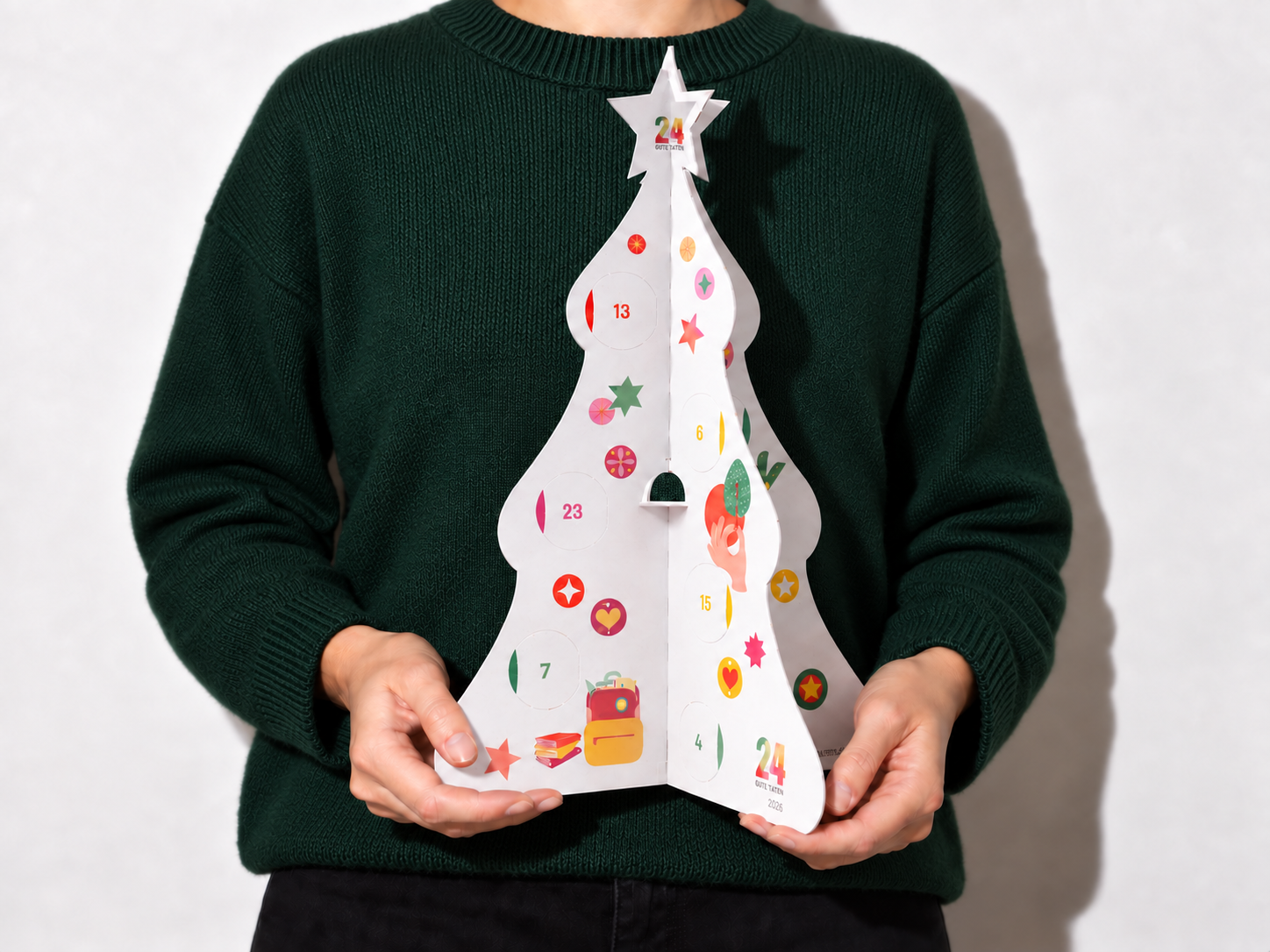 Der 24 GUTE TATEN Adventskalender 2026, gehalten von einer Person im grünen Pullover